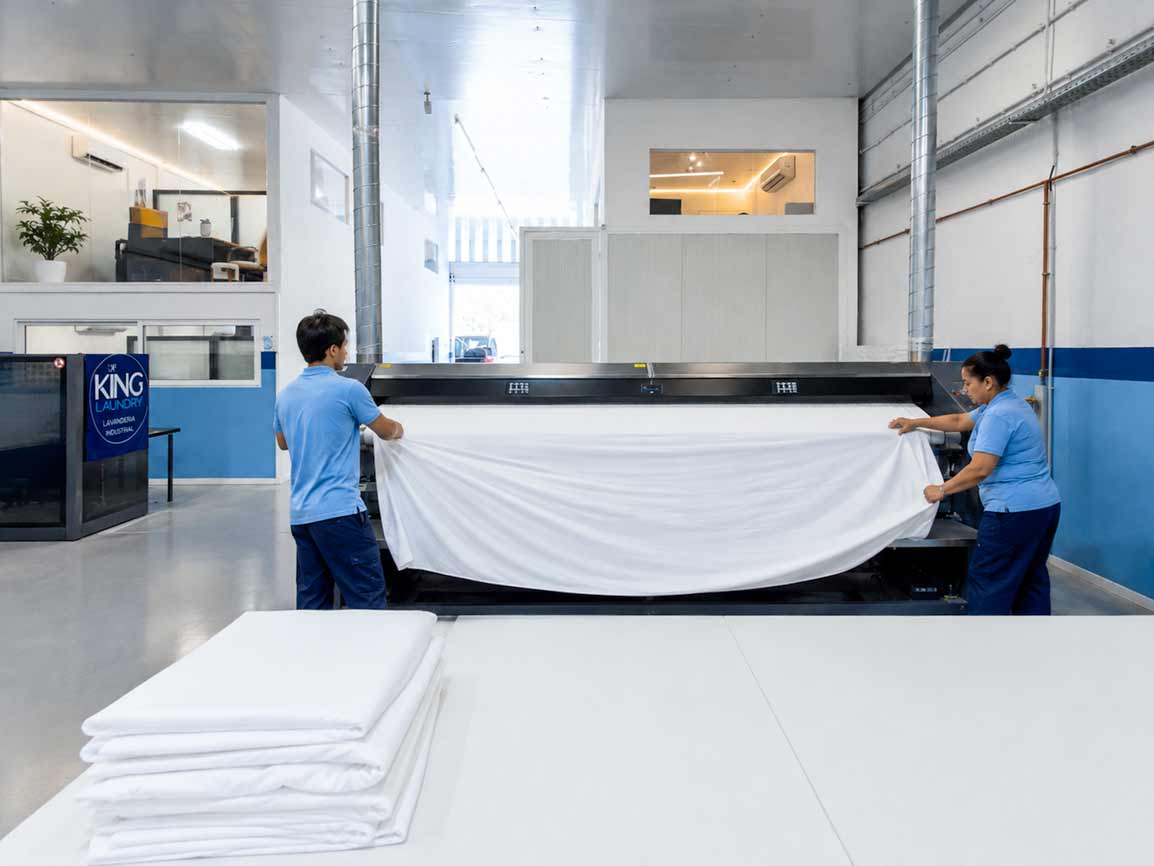 Two operators feeding a sheet into the ironer, with folded linen stacked in front.